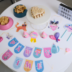 Table decorated for a party with a cat-themed banner spelling PARTY TIME, a white frosted cake on a stand, snack bowls, and cute cat and heart toppers.