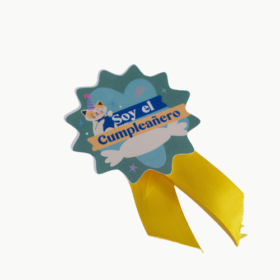Birthday badge with teal starburst, cartoon cat, and orange ribbon reading 'Soy el Cumpleañero' and yellow ribbon tails.
